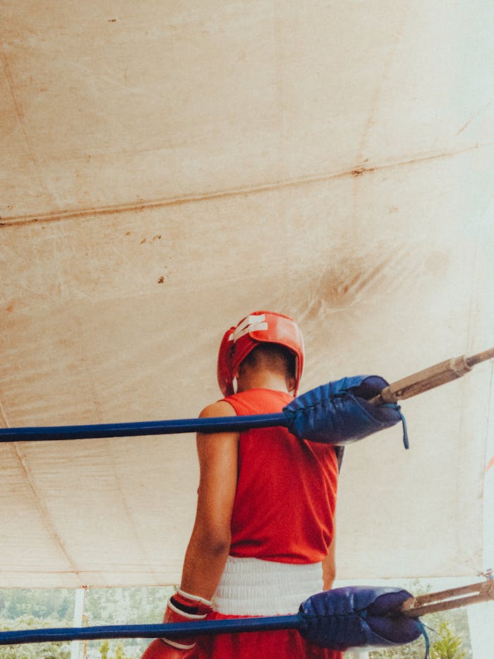 A boxer wearing red gear and helmet stands in the ring, viewed from behind.